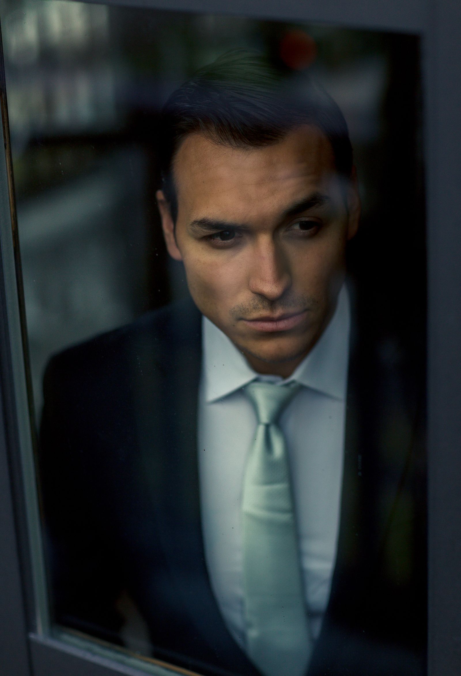A person seen through a window pane, looking down thoughtfully in soft daylight.