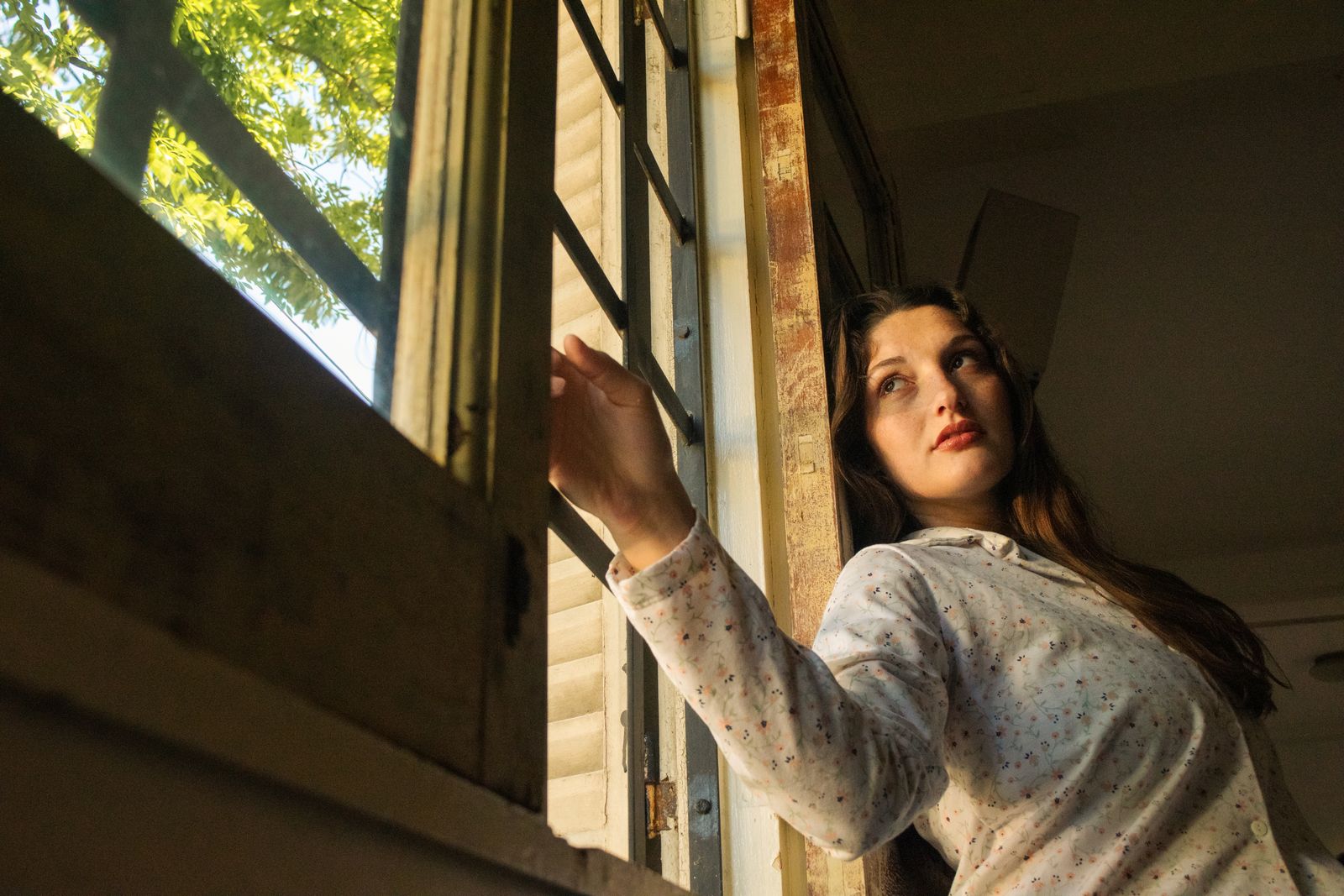 A young woman opening a window into warm sunlit foliage, quietly looking out.