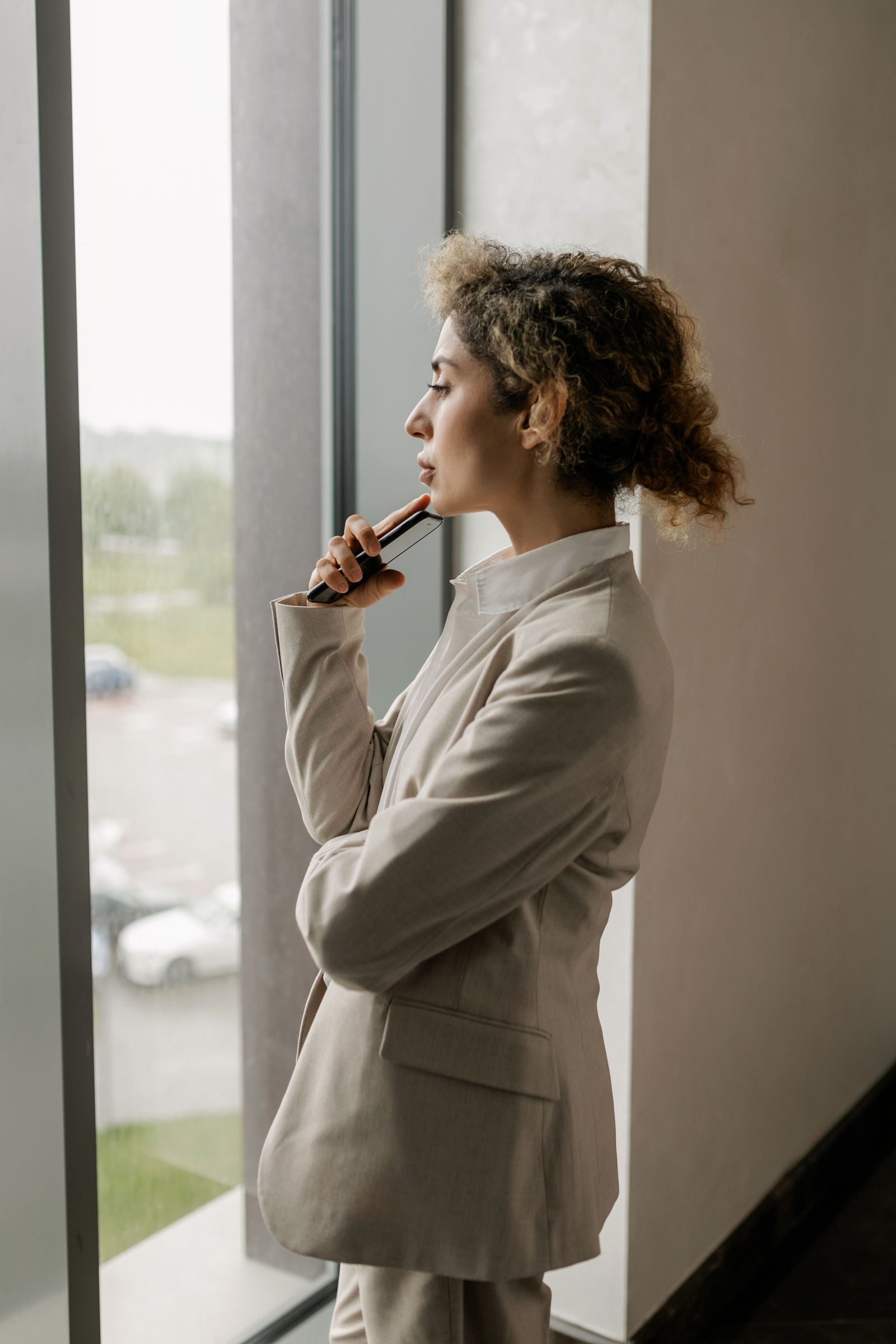 A professional in a beige suit standing quietly by an office window, holding her phone.