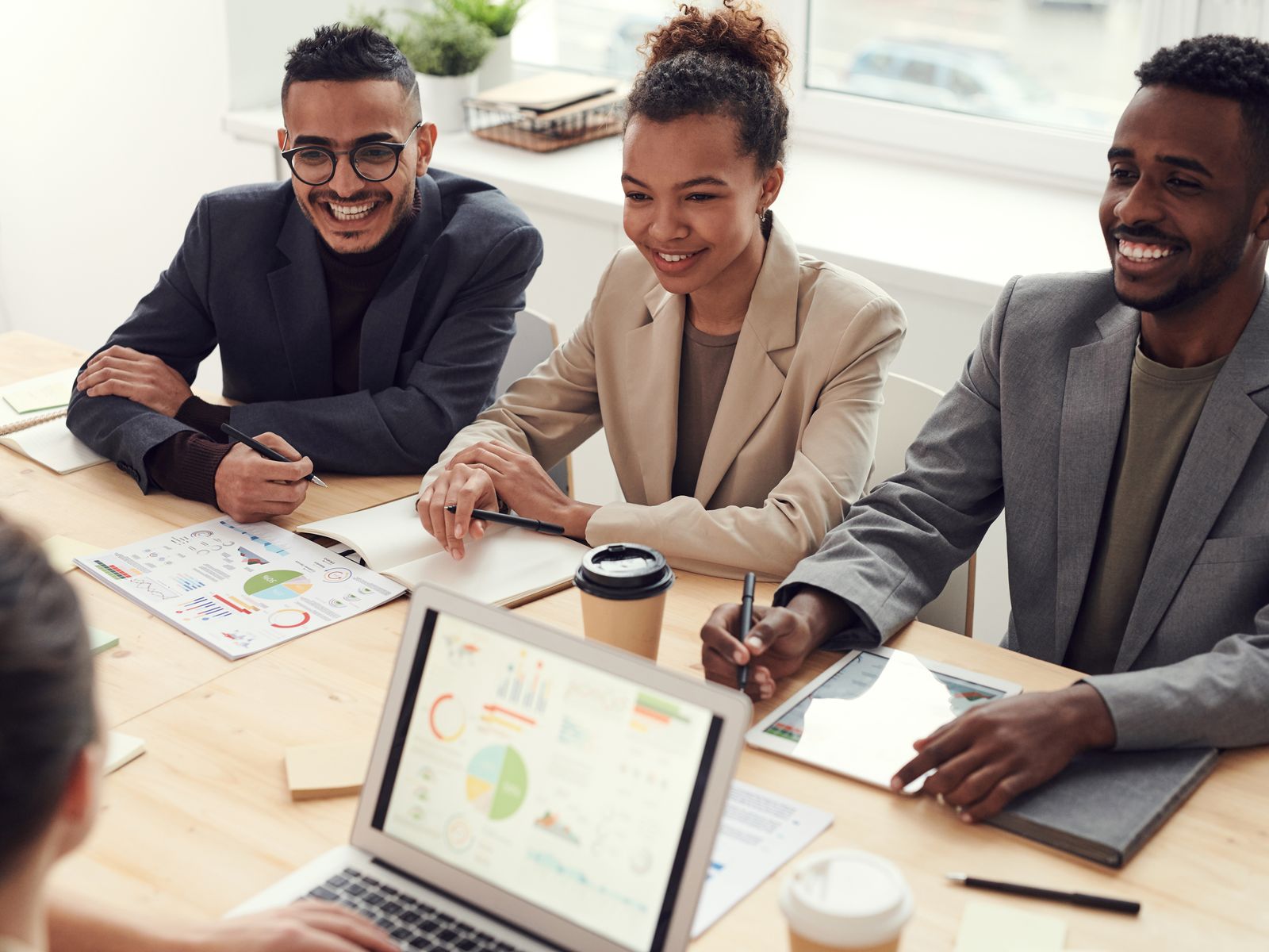 A diverse group of business professionals in a bright, modern workplace reviewing organizational reports and a laptop together during a working meeting.