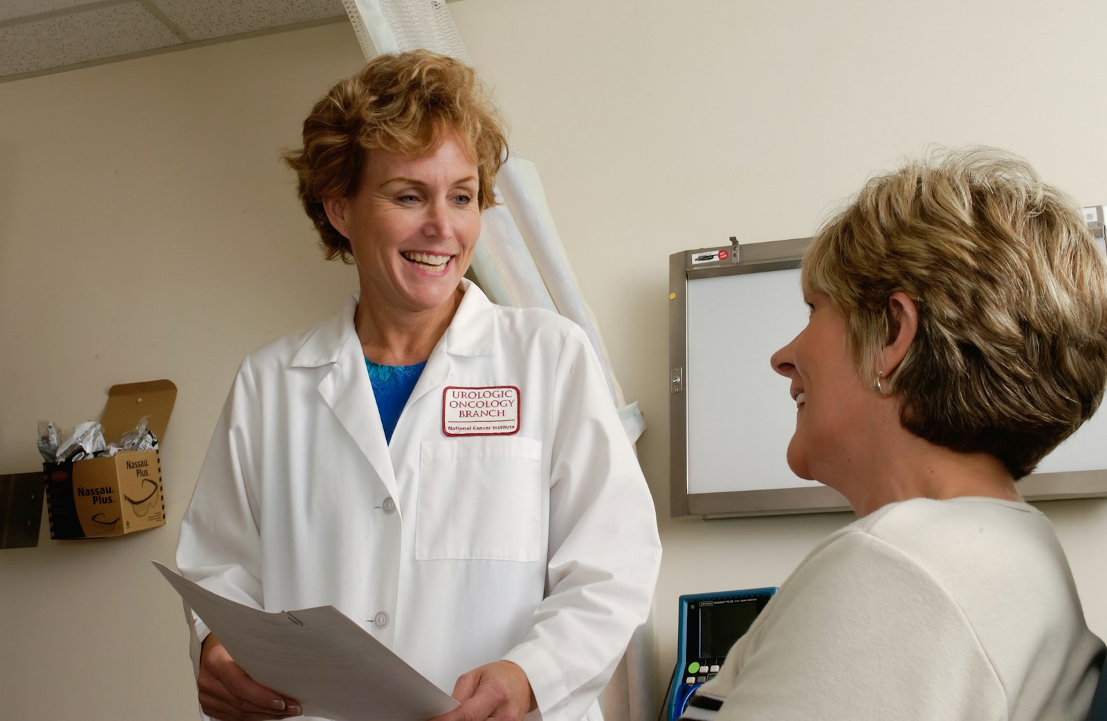 A clinician in a white lab coat in a warm, supportive conversation with a colleague inside a clinical office.