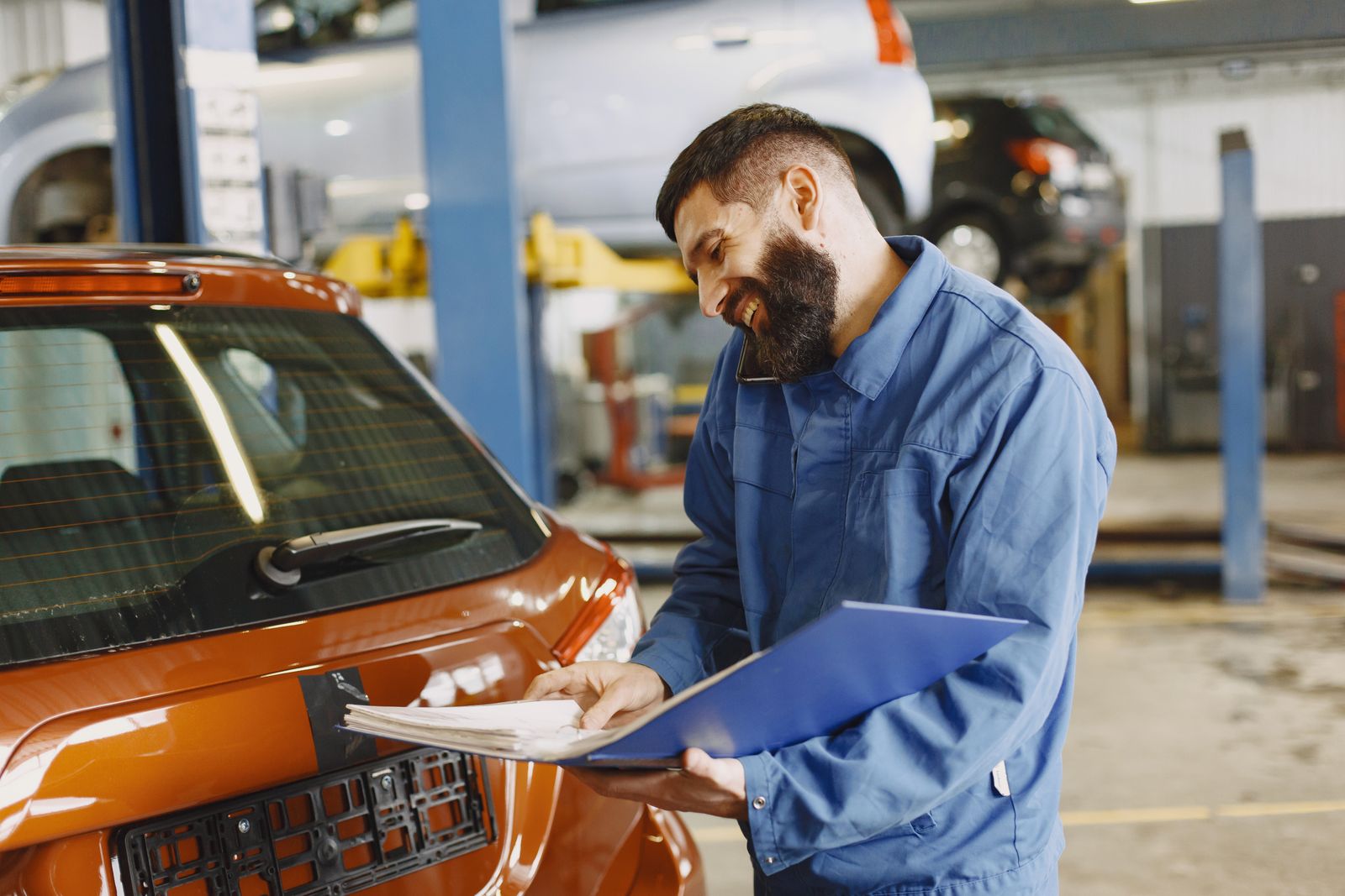 A smiling automotive service employee in coveralls with a clipboard beside a vehicle in a dealership service bay.