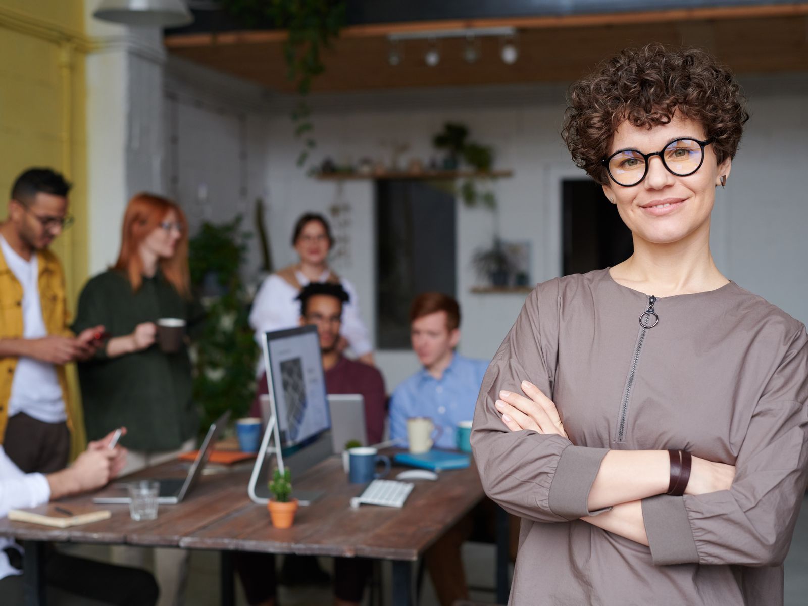 A confident brokerage team lead standing in a modern real estate office while colleagues collaborate at desks with laptops and computers behind her.