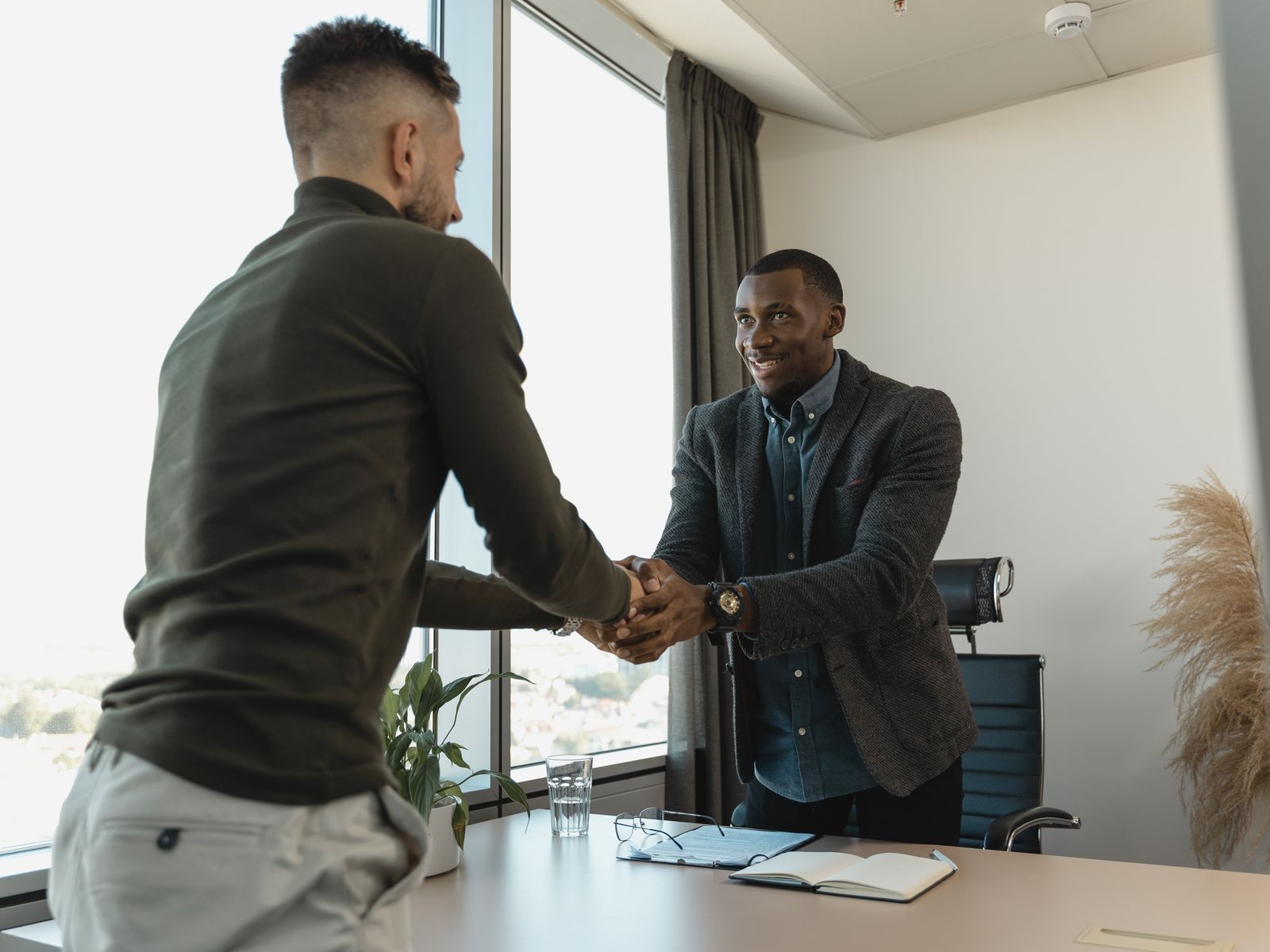 Two business professionals shaking hands across a desk in a modern real estate meeting room with contract paperwork visible.