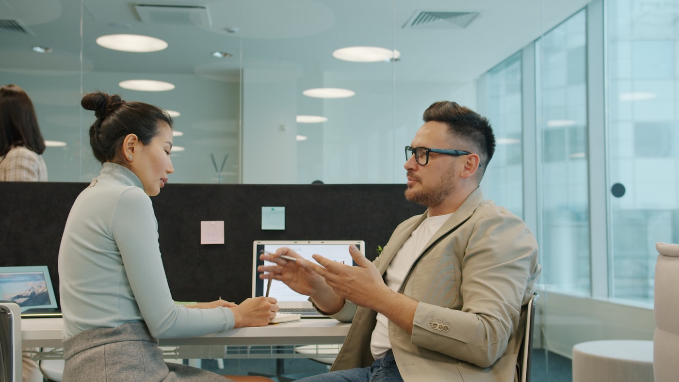A leader and a supportive professional engaged in a private, focused conversation at a desk in an office setting.